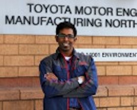Young man, dark skin, glasses, arms crossed, wearing dark blue jacket, standing in front of wall with words Toyota Motor Engineering.