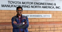 Young man, dark skin, glasses, arms crossed, wearing dark blue jacket, standing in front of wall with words Toyota Motor Engineering.