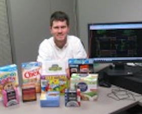 Young man, short dark hair, wearing white shirt, sitting at table covered with various boxes of toys and cereal.