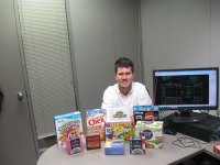 Young man, short dark hair, wearing white shirt, sitting at table covered with various boxes of toys and cereal.