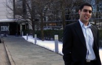 Young man, short dark hair, white shirt and dark suit coat, standing outside in front of building with IBM on side wall.