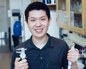 Young man, short dark hair, dark shirt, standing in lab room, holding pipette and medical instrument.