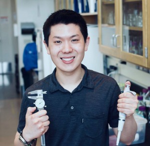 Young man, short dark hair, dark shirt, standing in lab room, holding pipette and medical instrument.