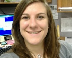 Young woman, long dark hair, grey sweater, silver short necklace, sitting in office in front of desk with back to computer display.