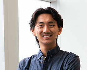 Young man, dark hair, dark blue shirt, standing with arms crossed over chest next to windows.