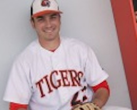 Young man, in baseball uniform, cap, wearing mitt, shirt says Tigers, shelf on wall with athletic shoes.