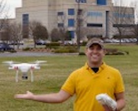 Young man wearing yellow polo shirt, green cap, standing outside in lawn, hand indicating hovering drone, building in background with letters UAH.