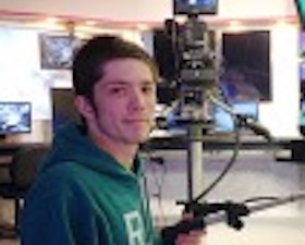 Young man, short brown hair, green hoodie, hand on large video camera control.