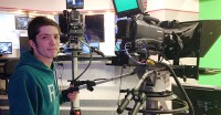 Young man, short brown hair, green hoodie, hand on large video camera control.
