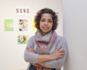 Young woman, short dark hair, arms crossed, grey top and multi-colored scarf around neck, standing in front of wall with graphic images.