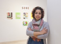 Young woman, short dark hair, arms crossed, grey top and multi-colored scarf around neck, standing in front of wall with graphic images.