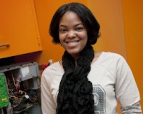 Young woman, dark skin, white jersey top, dark pants, dark long knitted scarf around neck, standing by computer open to show circuitry.