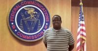 Young man, dark skin, glasses, striped shirt, hands crossed behind back, standing in front of  Federal Communications Commission plaque and American flag.