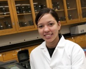 Young woman, dark hair tied back, wearing white lab coat, blue lab gloves, leaning elbow on lab table.