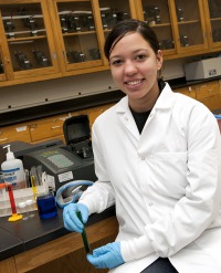 Young woman, dark hair tied back, wearing white lab coat, blue lab gloves, leaning elbow on lab table.