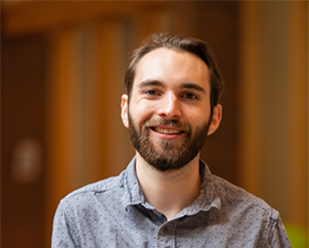 Photo of young man with dark hair and beard, smiling, wearing light blue shirt and leaning on round stair banister.