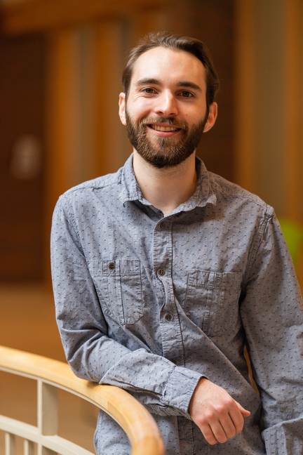 Photo of young man with dark hair and beard, smiling, wearing light blue shirt and leaning on round stair banister.