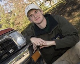 Young man, dark hoodie, tan pants, tan baseball cap, kneeling down in front of V6 vehicle with Wyoming license plate.