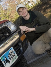 Young man, dark hoodie, tan pants, tan baseball cap, kneeling down in front of V6 vehicle with Wyoming license plate.