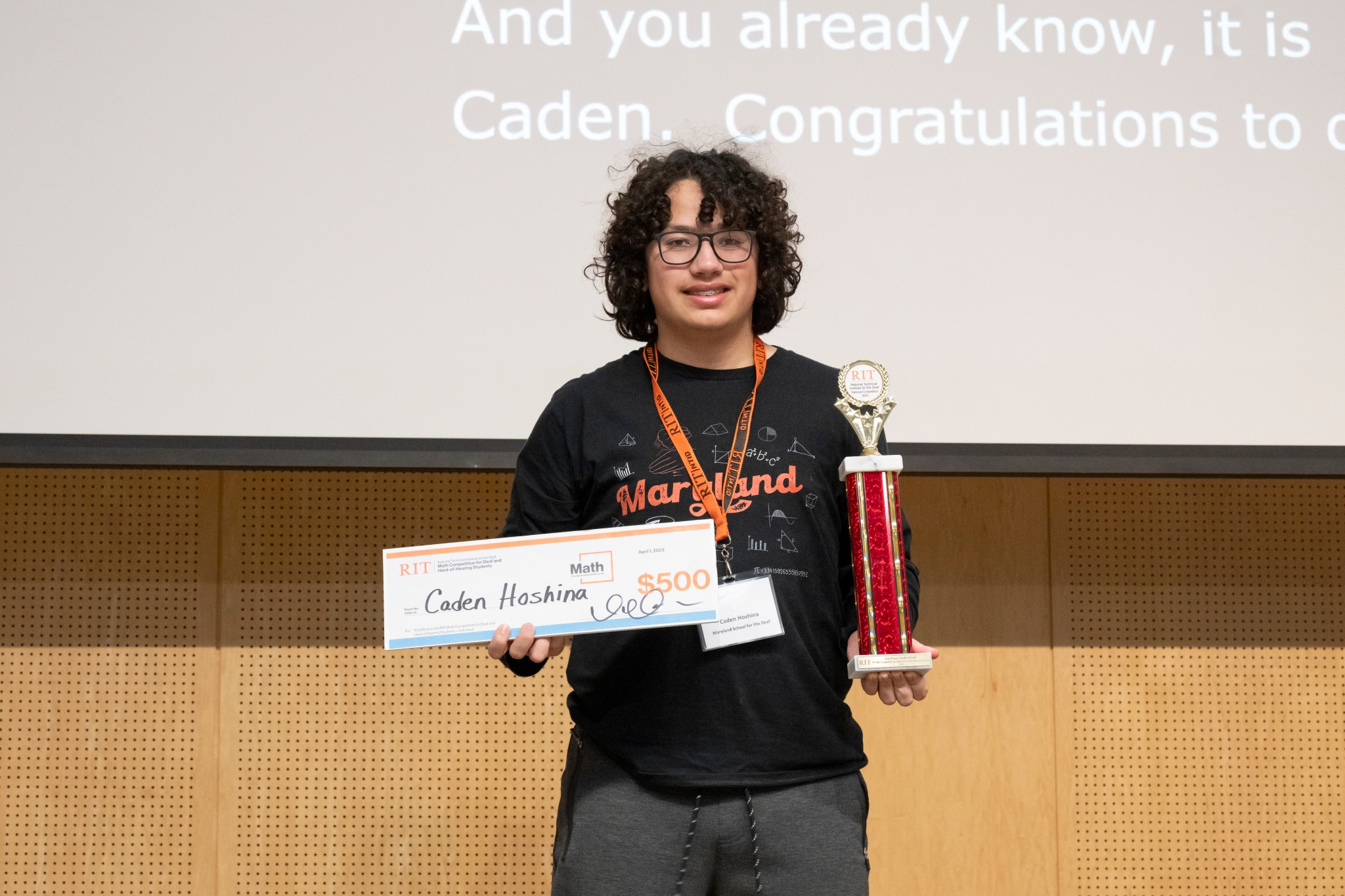 A middle school contestant from Maryland School for the Deaf stands facing the camera, holding a trophy and a check for $500. The contestant is wearing a black and orange shirt with an orange name tag.