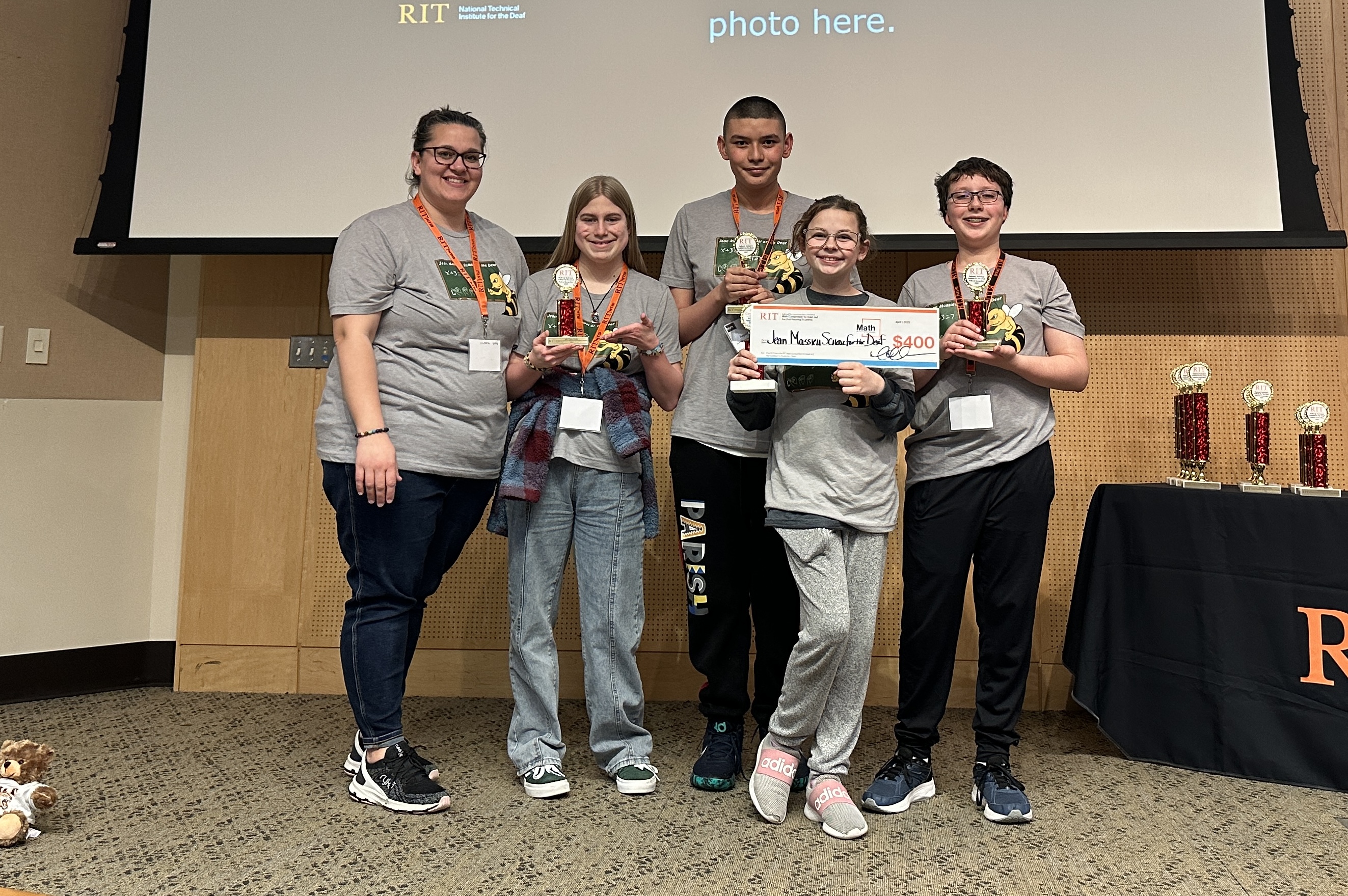The image shows Jean Massieu School for the Deaf contestants and their coach in Grey t-shirts, standing in line, facing the camera and holding up trophies and a check for $400. Each person is wearing an orange name tag.