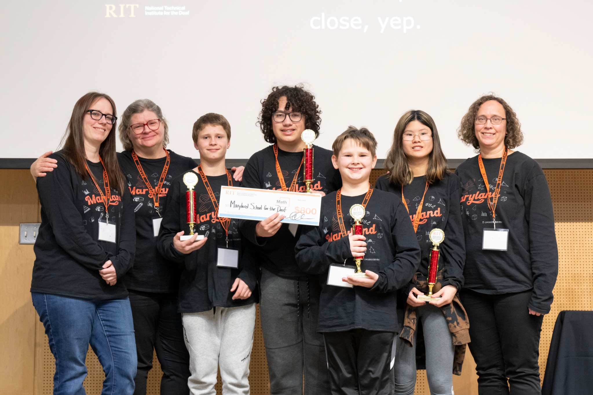 The image shows Maryland School for the Deaf contestants and their coaches in black and orange team shirts, standing in line, facing the camera and holding up trophies and a check for $800. Each person is wearing an orange name tag. 