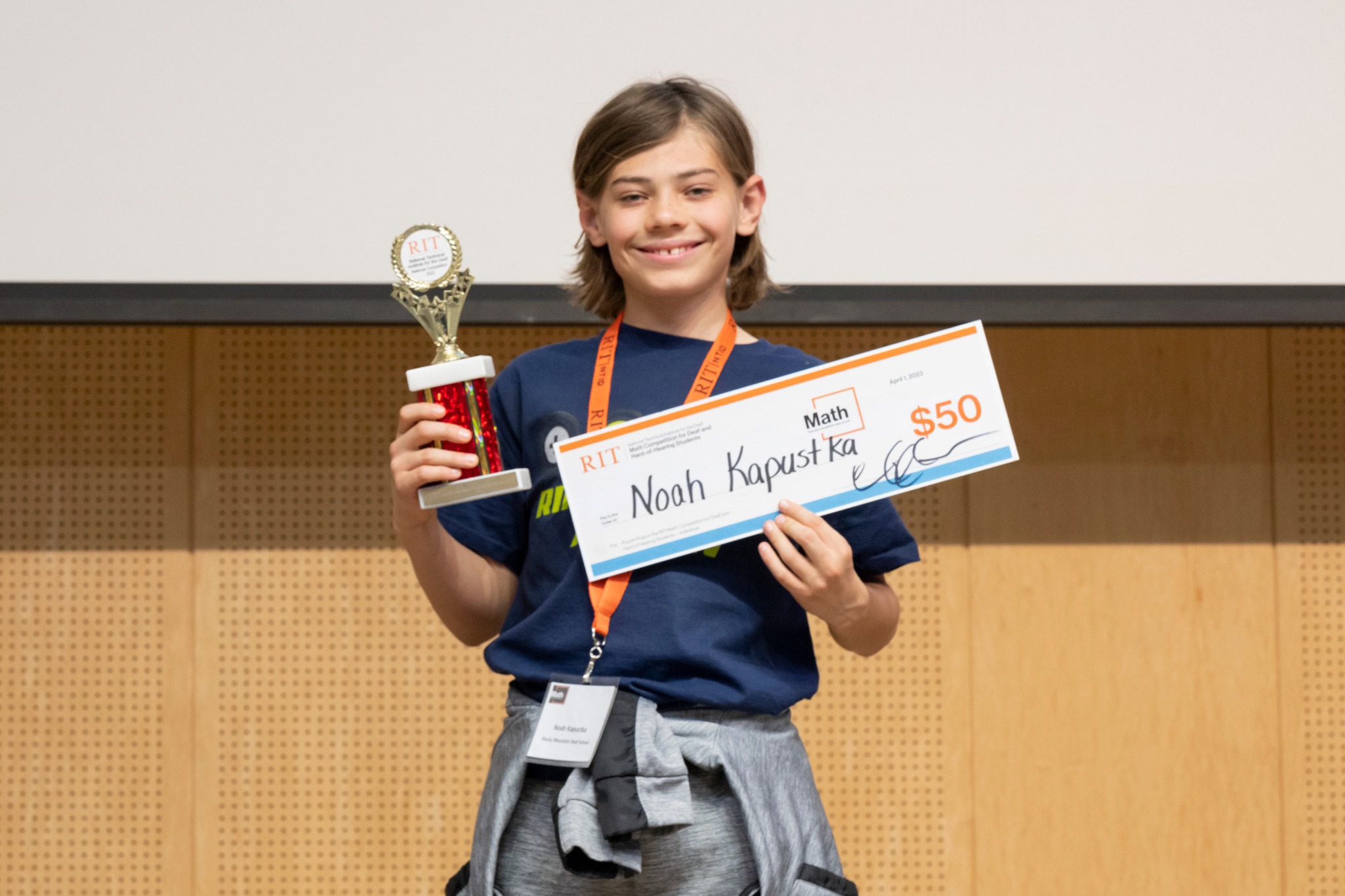A middle school contestant from Rocky Mountain Deaf School stands facing the camera, holding a trophy and a check for $50. The contestant is wearing a blue shirt with an orange name tag.