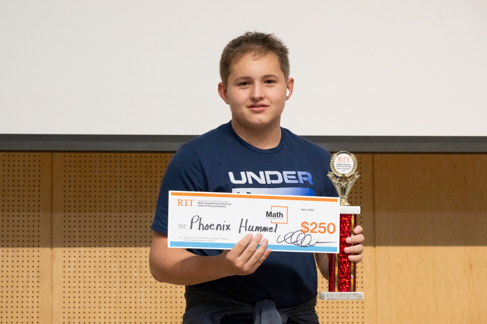 A middle school contestant from Texas School for the Deaf stands facing the camera, holding a trophy and a check for $250. The contestant is wearing a blue shirt.