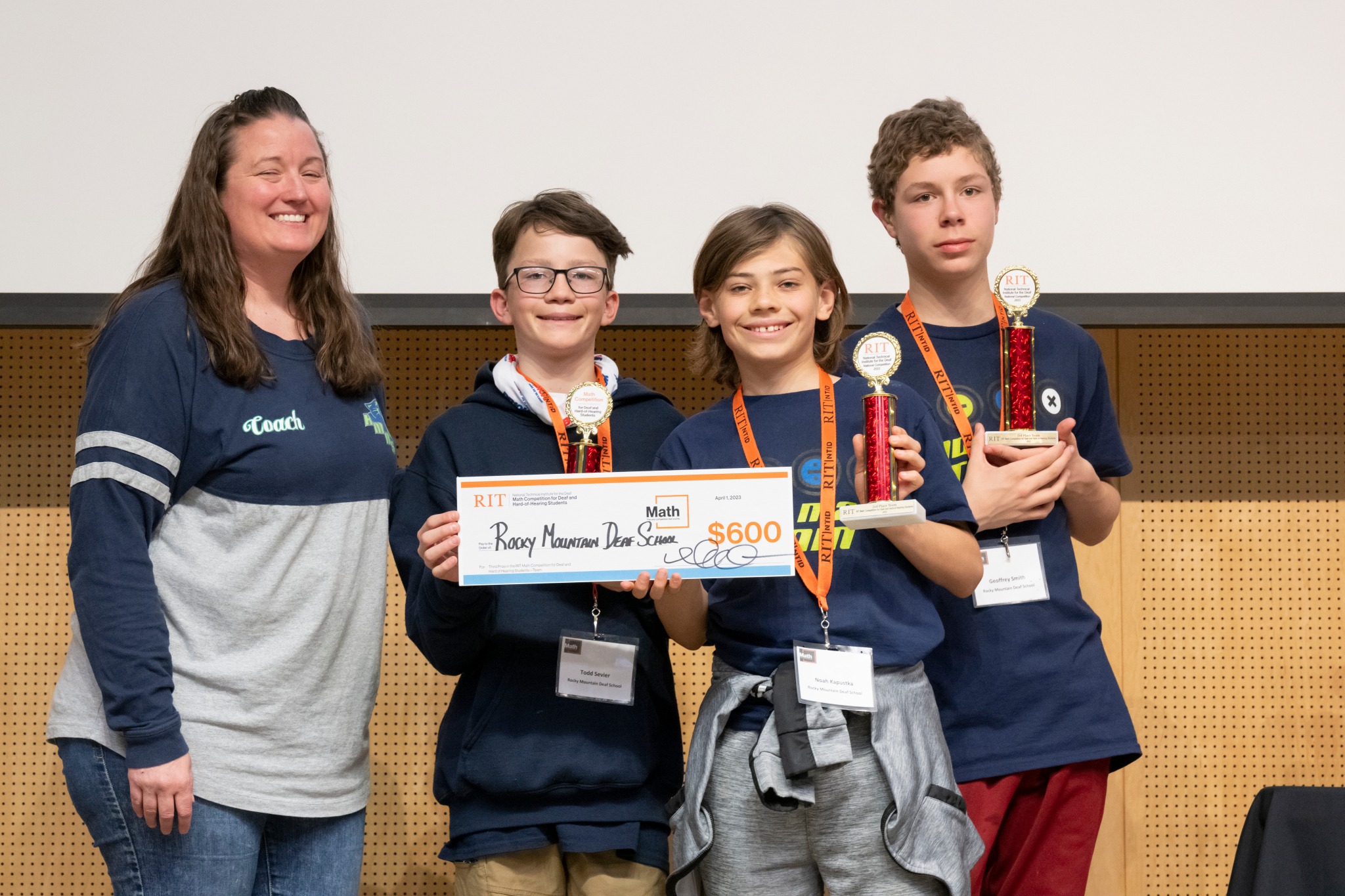 The image shows Rocky Mountain Deaf School contestants and their coach in blue and grey t shirts, standing in line, facing the camera and holding up trophies and a check for $600. Contestants are wearing orange name tags. 