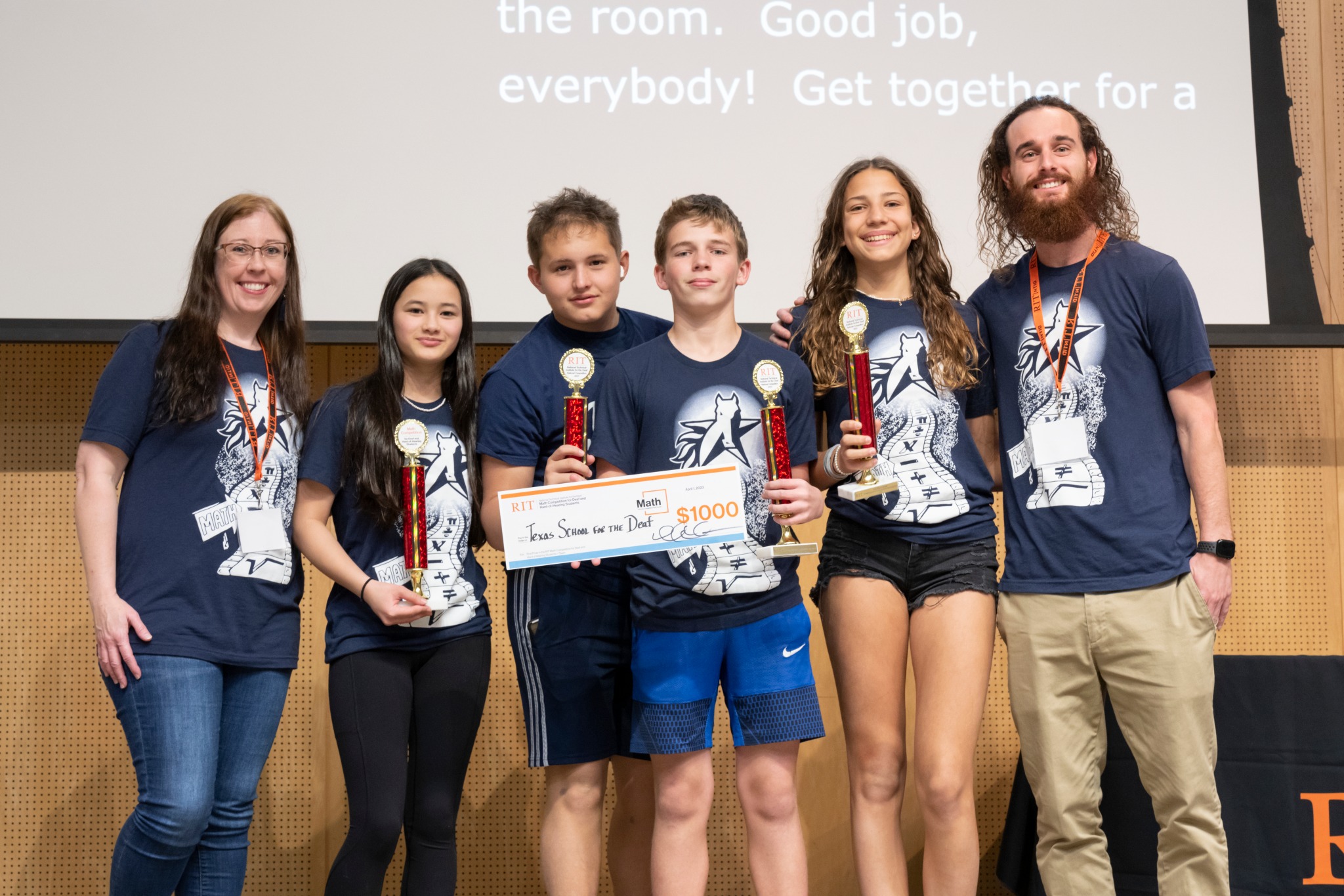The image shows Texas School for the Deaf contestants and their coaches in blue team shirts, standing in line, facing the camera and holding up trophies and a check for $1,000.