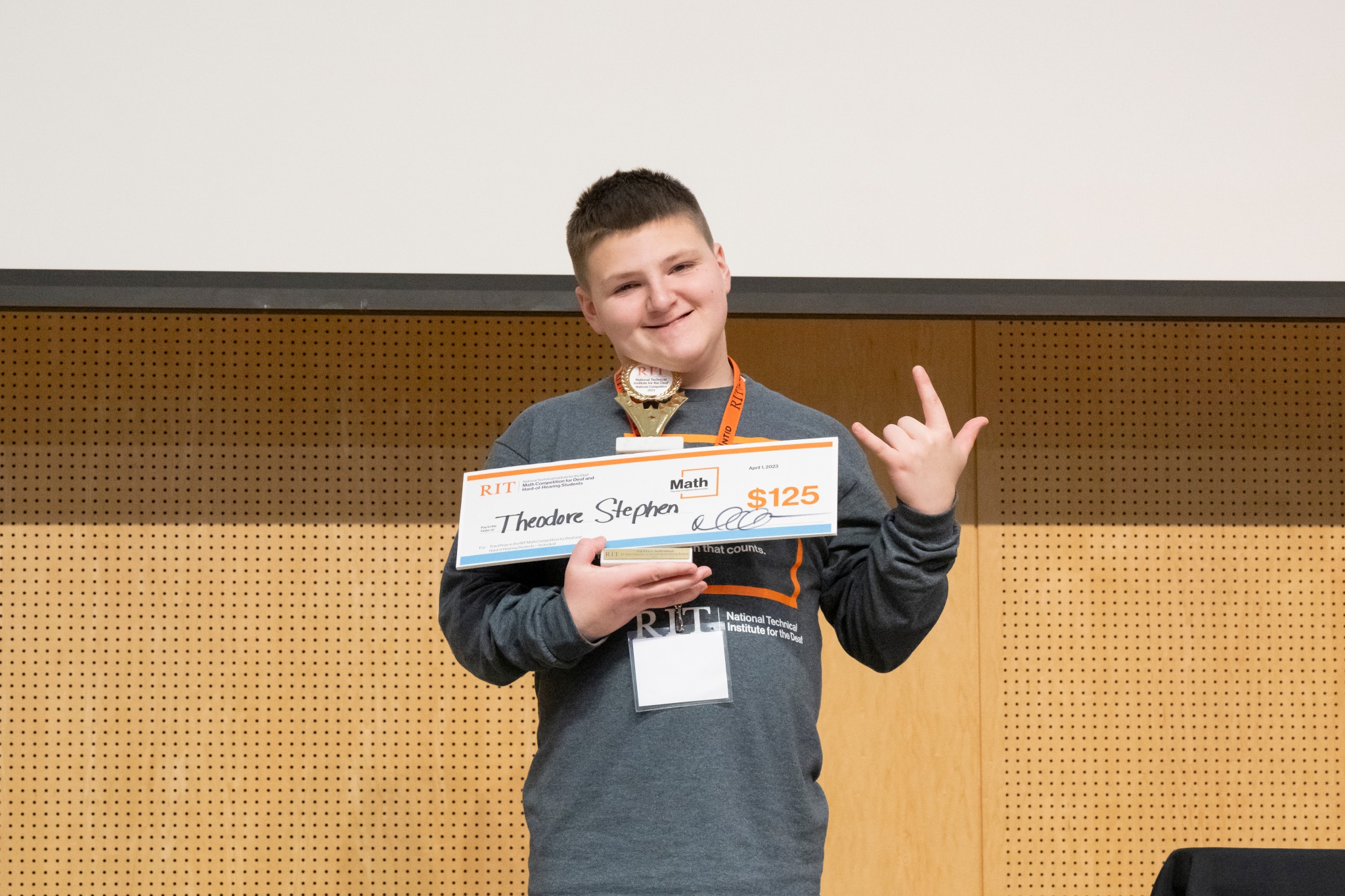 A middle school contestant from Delaware School for the  Deaf stands facing the camera, holding a trophy and a check for $125. The contestant is wearing a grey RIT Math Competition shirt with an orange name tag.