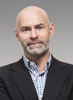Photo of man, bald, dark suit jacket, striped shirt open at color.