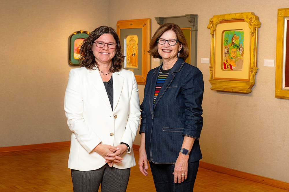 Two individuals standing in an art gallery with framed paintings behind them, one wearing a white blazer and glasses, the other in a dark blazer and glasses.