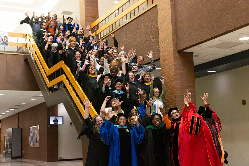 Large group of people posing for a photo on stairs indoors, with hands waving.