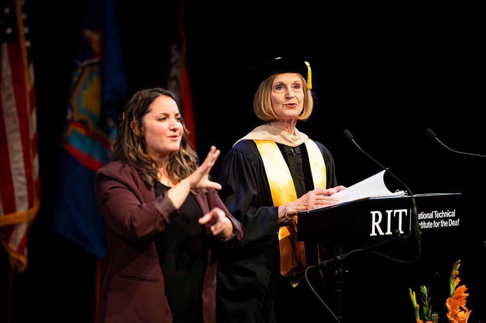Person speaking at a podium with sign language interpreter, other individuals seated on stage.