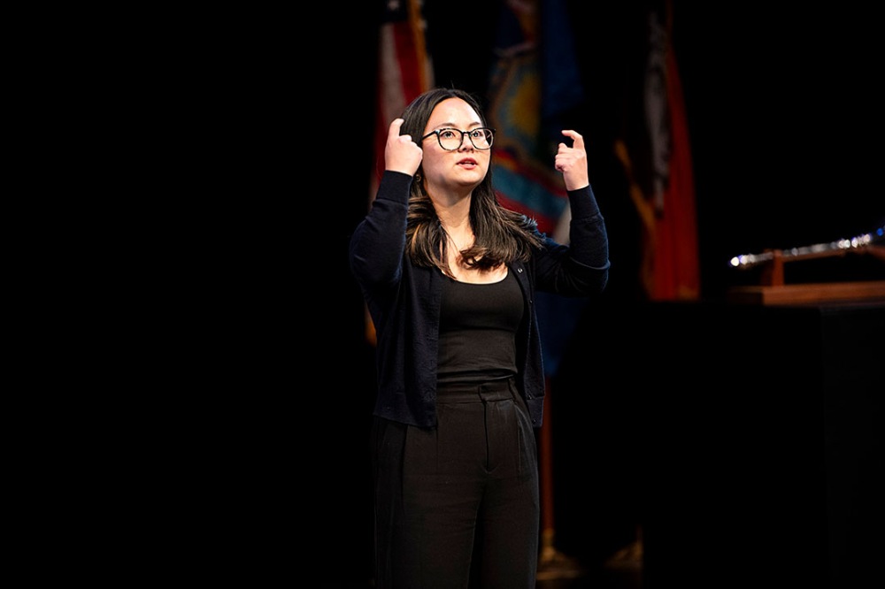 Individual signing with hands raised, flags visible in the background