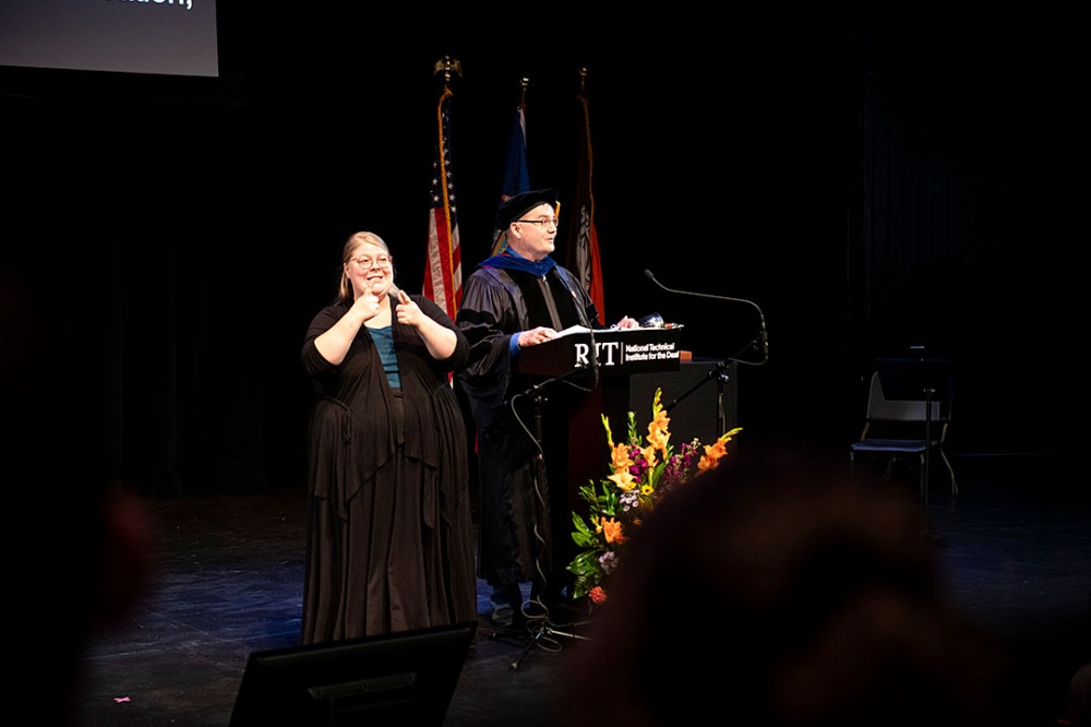 Person speaking at podium with sign language interpreter next to them, flags and flowers visible in the background.