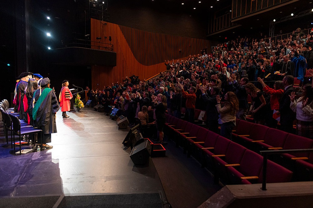 Audience applauding while individuals in academic regalia stand on stage.