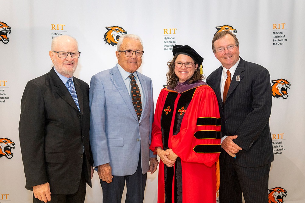 Four individuals standing together in front of an RIT backdrop, with one person wearing academic regalia.