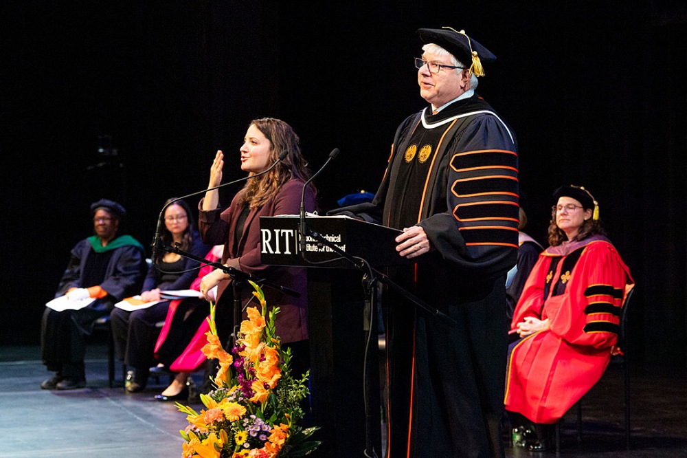 Person speaking at a podium with sign language interpreter, other individuals seated on stage.