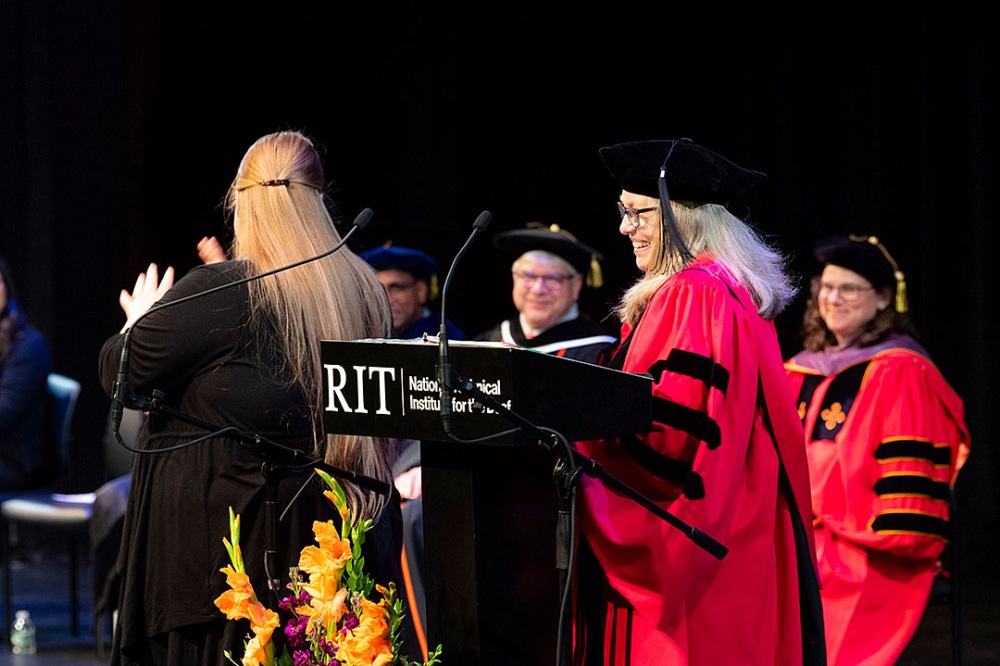 Person in red academic regalia smiling at the podium, another person signing next to them.