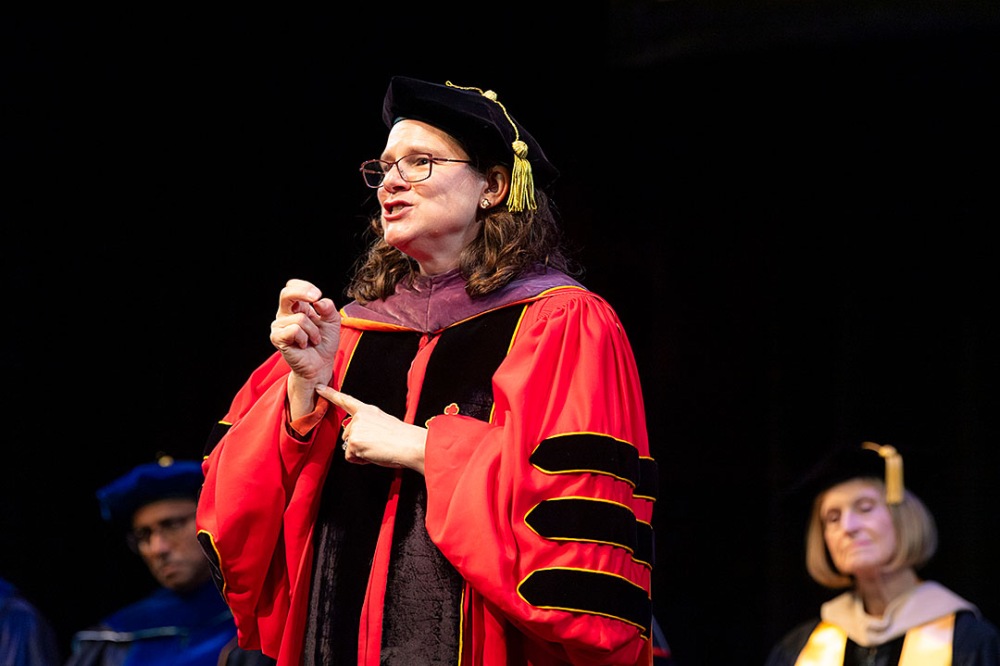 Individual wearing red academic regalia signing while standing on stage.