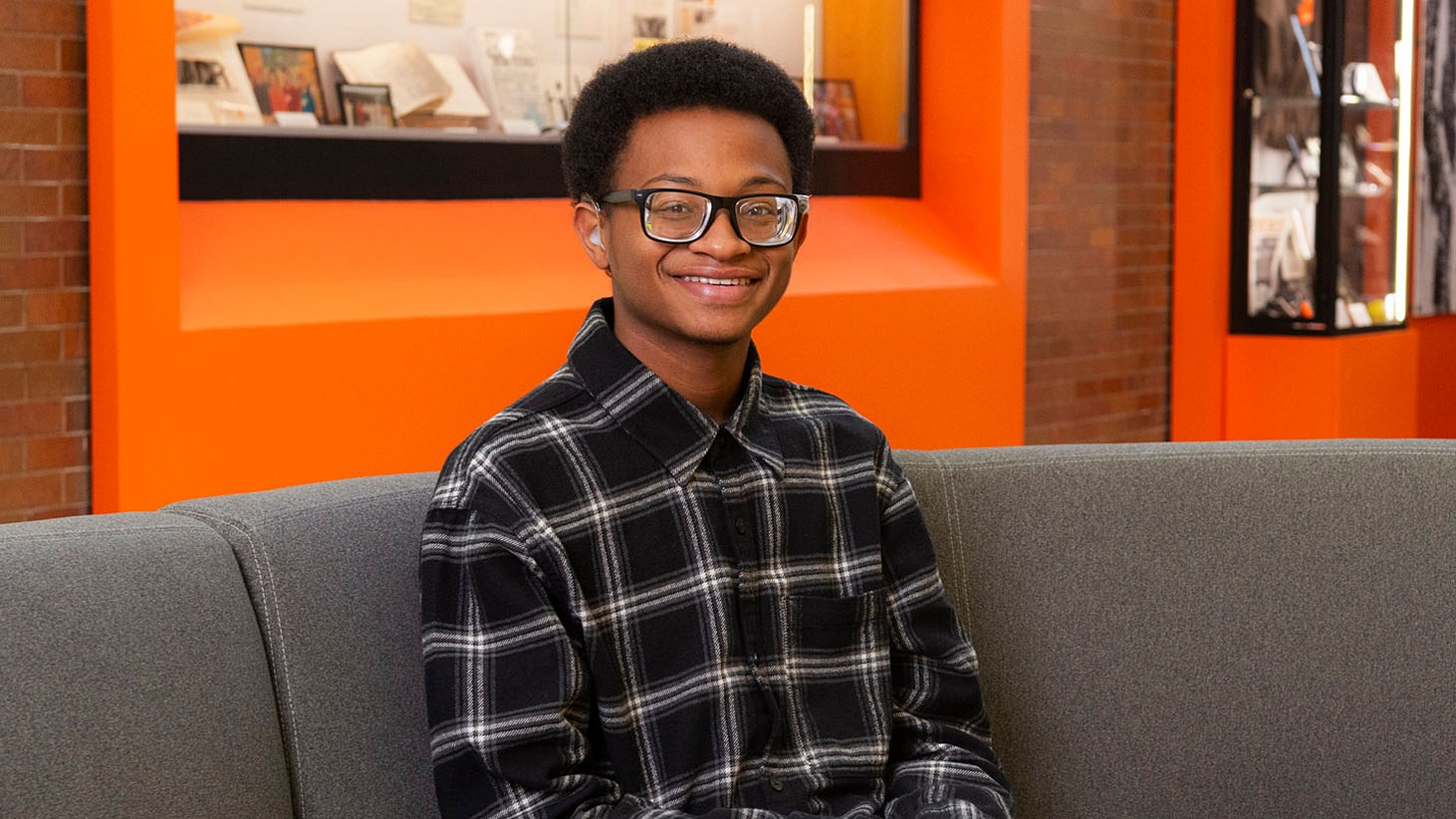 Person seated on a gray couch in a brightly lit space with an orange wall, wearing a black-and-white plaid shirt.