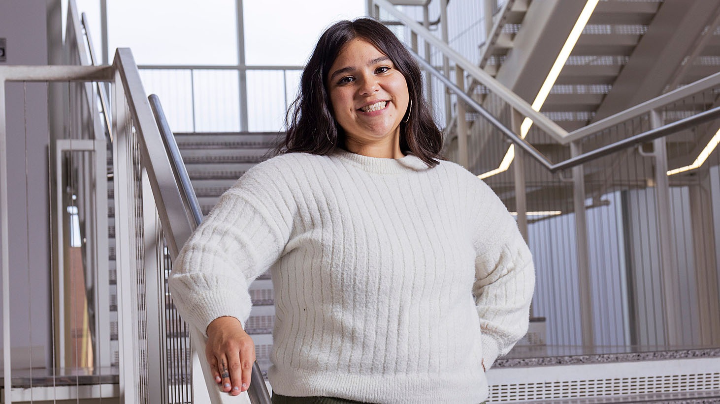 Person standing on an indoor staircase wearing a white knit sweater, with one hand resting on the railing.