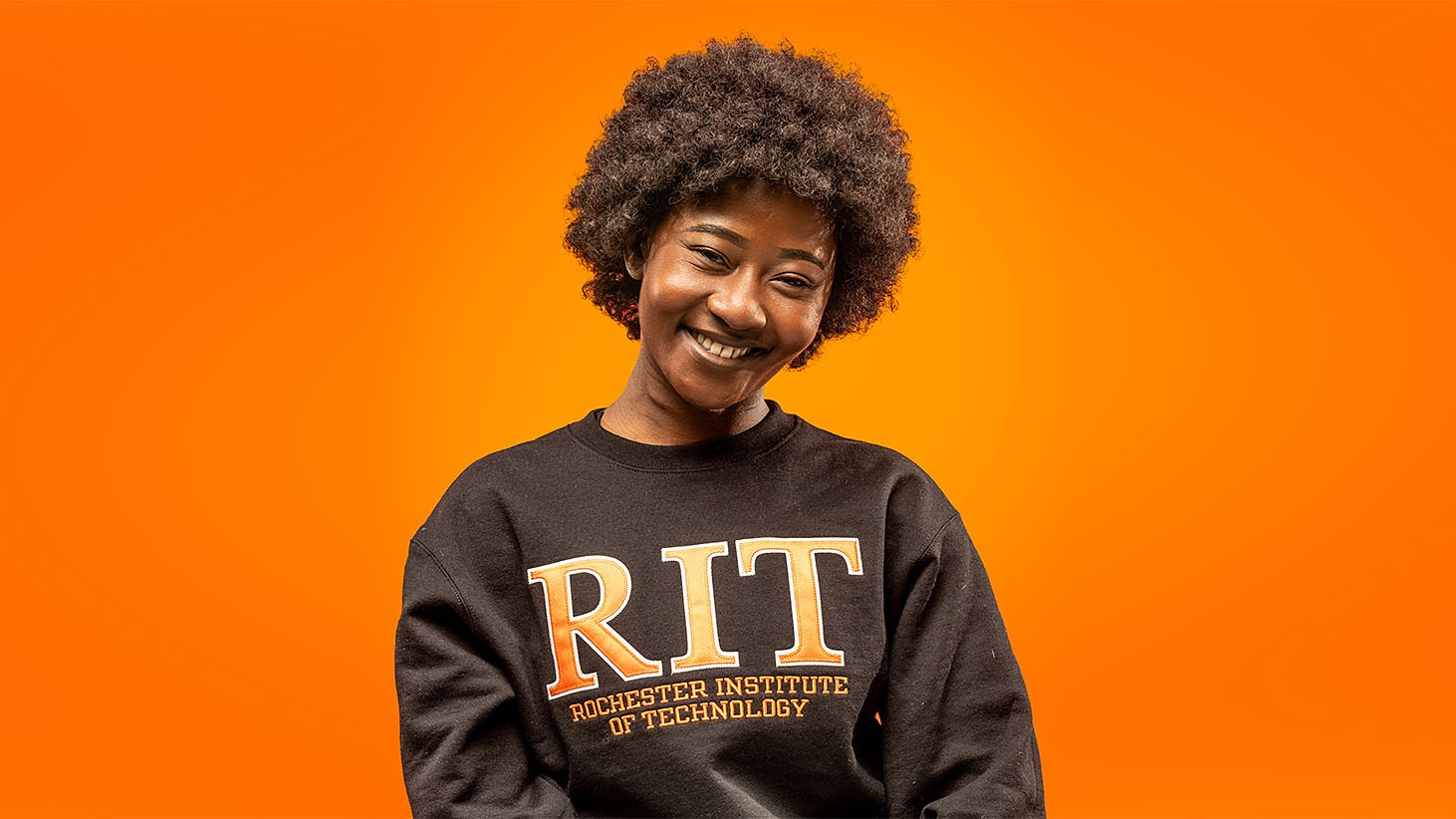 Person seated against an orange background wearing a black sweatshirt with “RIT Rochester Institute of Technology” printed on it.