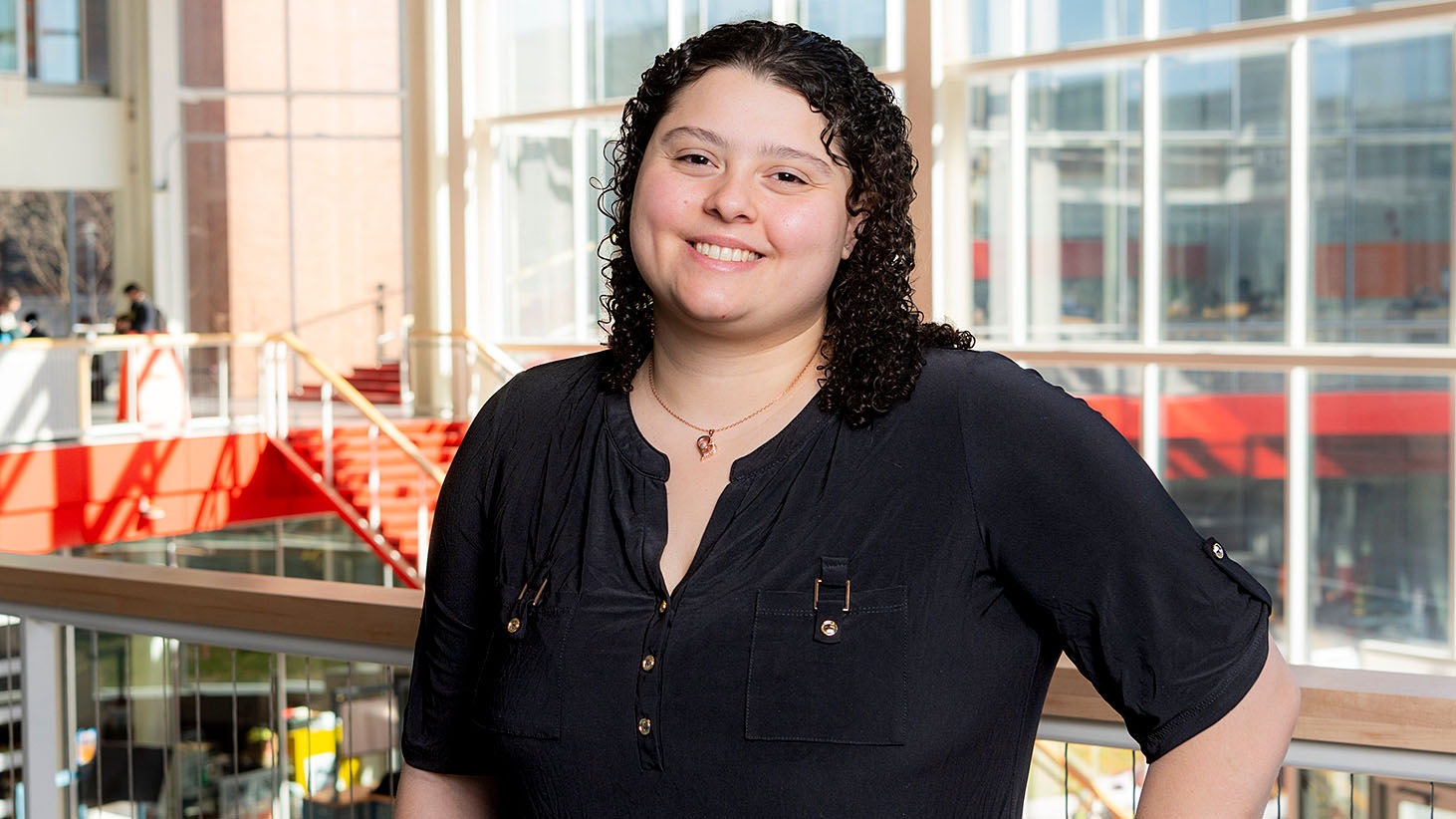 Person standing inside a modern building with large windows, wearing a navy blouse with button details.