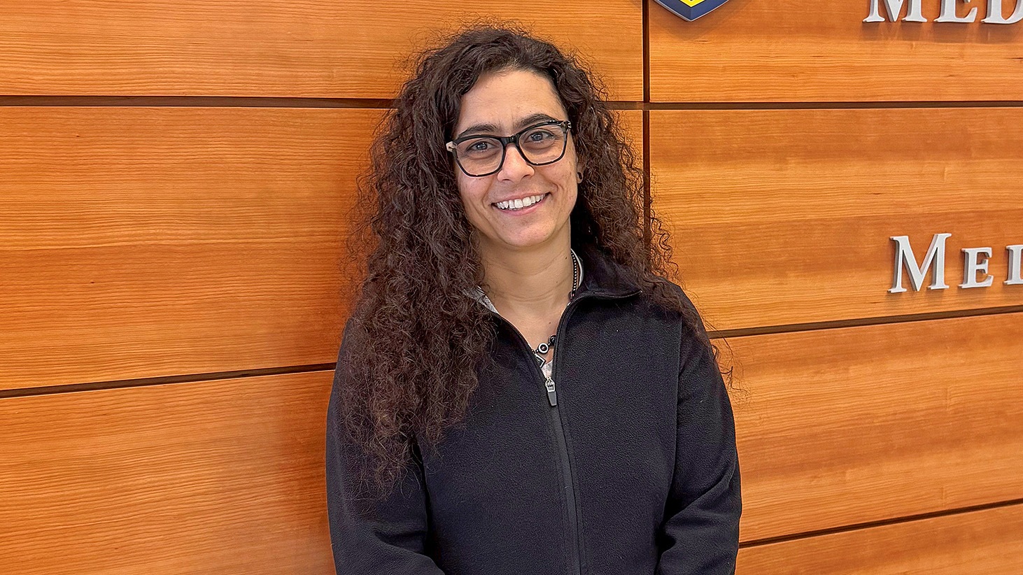 A smiling woman with long curly dark hair and black-rimmed glasses, wearing a black zip-up fleece jacket. She is standing indoors in front of a wall made of horizontal wooden panels.