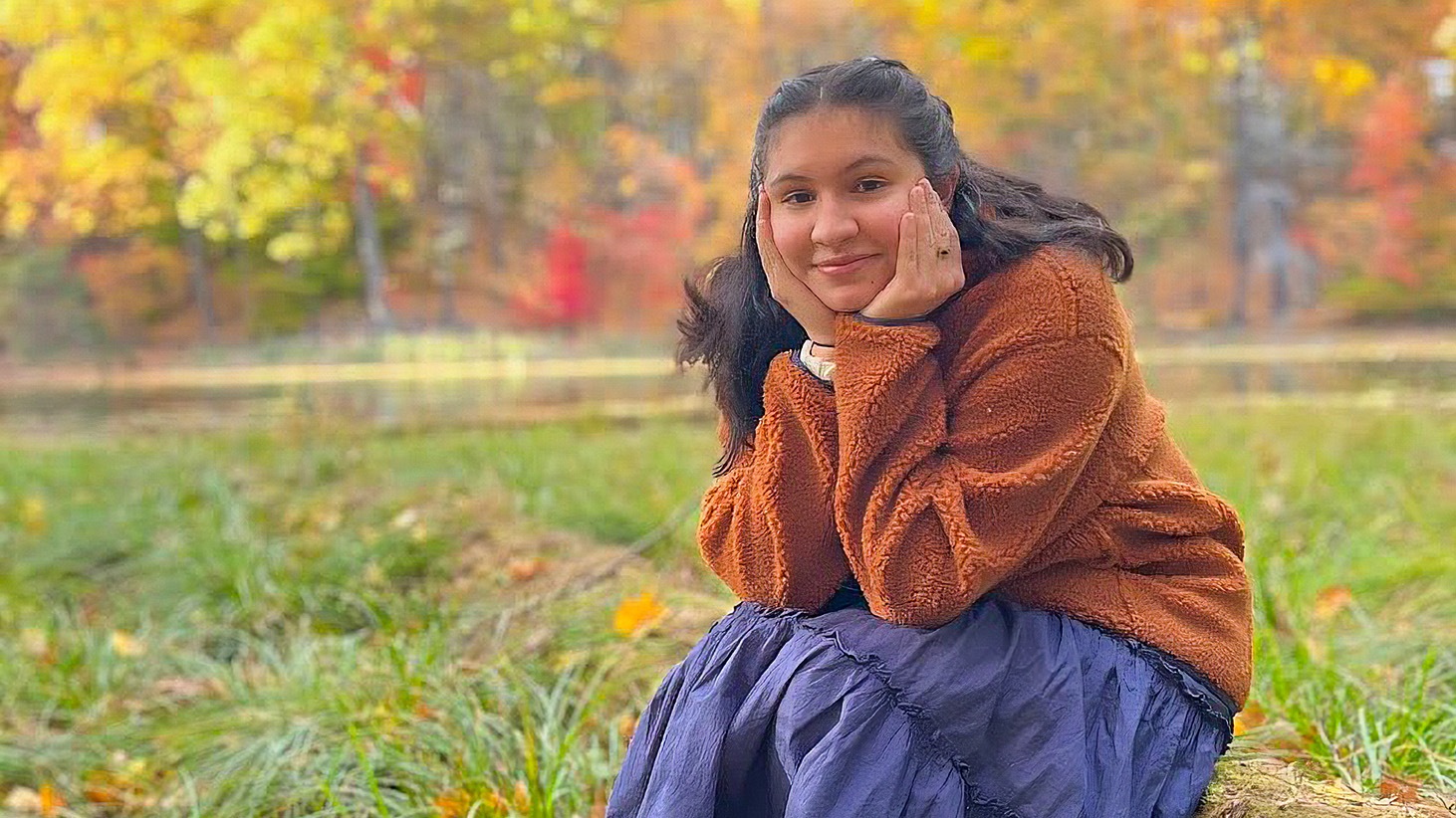 A young woman with long dark hair smiling while sitting outdoors, resting her face in both hands. She is wearing a fuzzy orange pullover and a long dark blue skirt. The background features green grass, a body of water, and trees with vibrant autumn foliage.