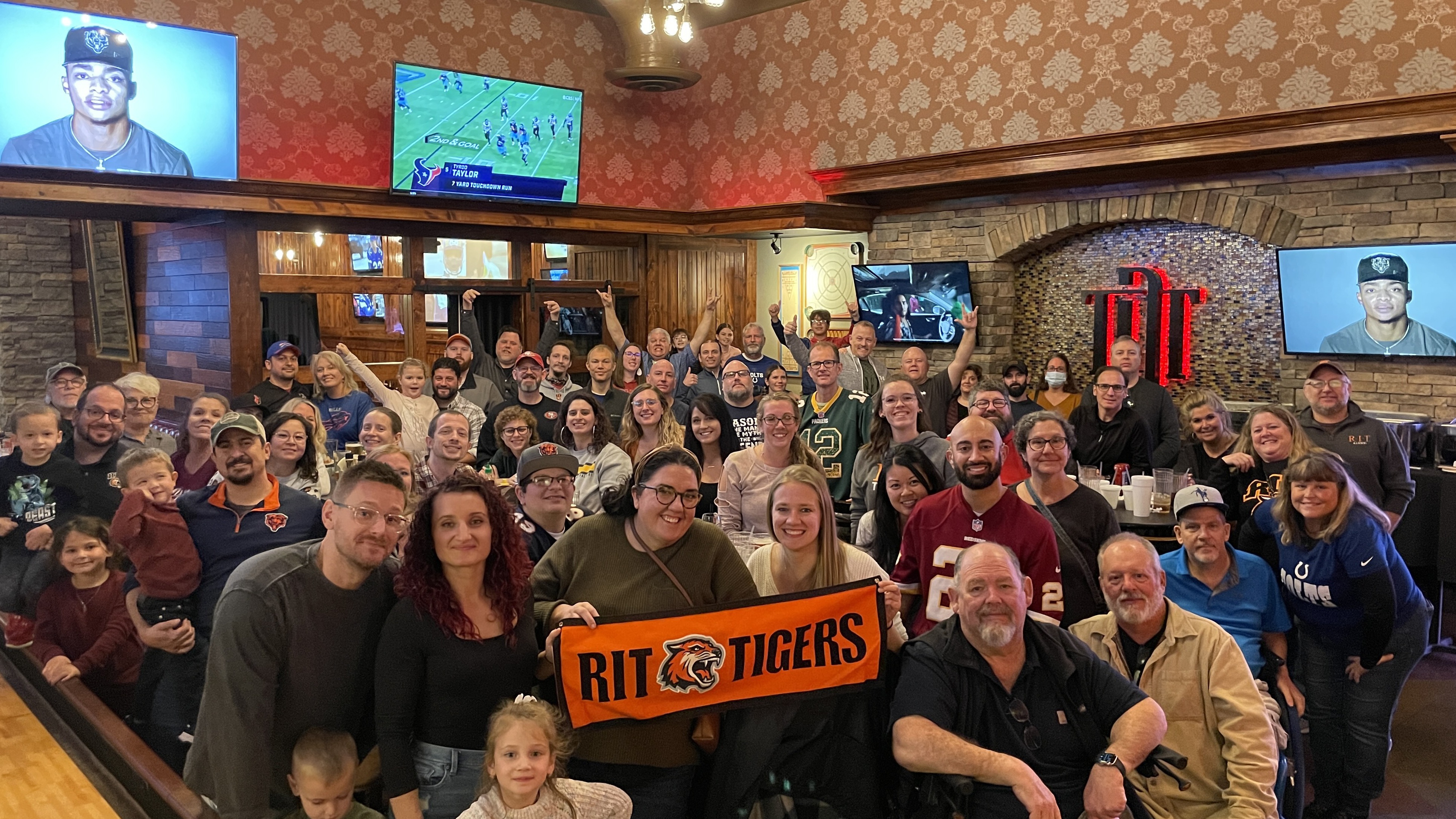 Photo of a large group of people in a sports bar. Two people in front are holding an orange banner that reads "RIT Tigers."