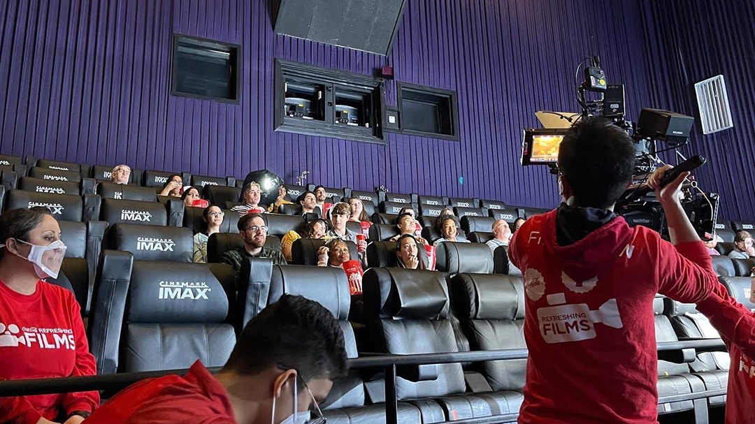Students sitting in theater eating popcorn while being filmed. 
